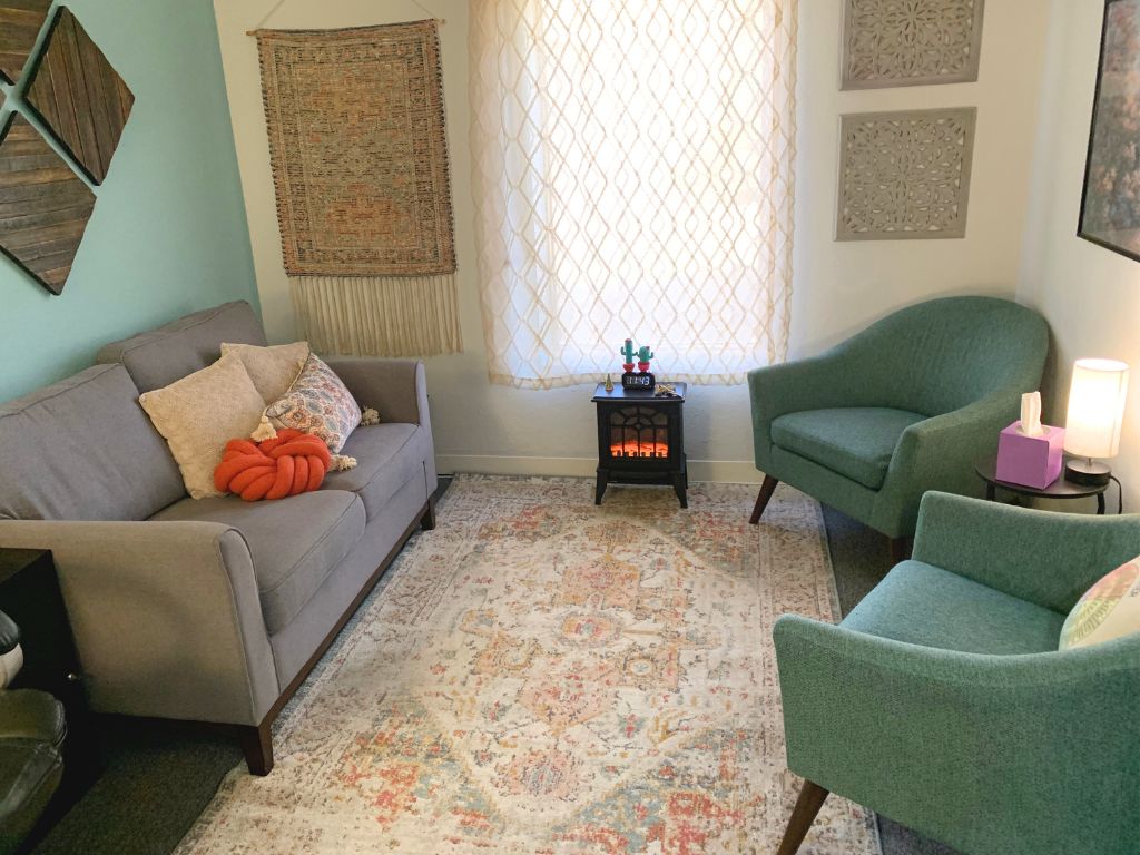 Office space for therapy with blue walls, cream rug, a grey couch, and two blue chairs in front of an electric fireplace and window